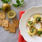 kiwi fruit and cheese on top of whole grain cracker with white circle platter and marble background