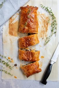 vegetable wellington on parchment paper next to chef knife and herb sprigs