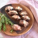 blue cheese and rosemary stuffed dates wrapped in prosciutto on a brown wood plate rosemary sprig and red and white towel