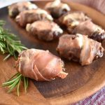 blue cheese and rosemary stuffed dates wrapped in prosciutto on a brown wood plate, rosemary sprig and red and white towel