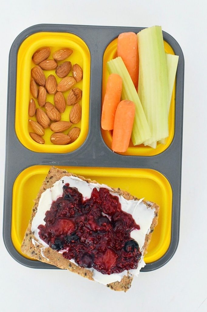 yellow bento box with almonds, carrots, celery sticks and mixed berry chia jam and mascarpone cheese on crispbread