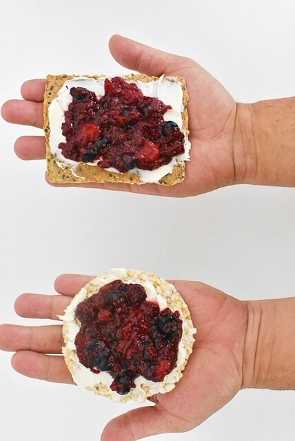2 hands holding crispbread and brown rice cake topped with mixed berry chia jam and mascarpone cheese