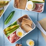 white wood background with breakfast bento box containing sliced Persian cucumber, sliced pear, crispbread, hard boiled eggs, prosciutto slices and unsalted almonds
