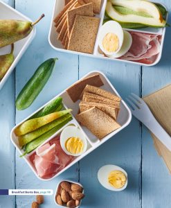 white wood background with breakfast bento box containing sliced Persian cucumber, sliced pear, crispbread, hard boiled eggs, prosciutto slices and unsalted almonds